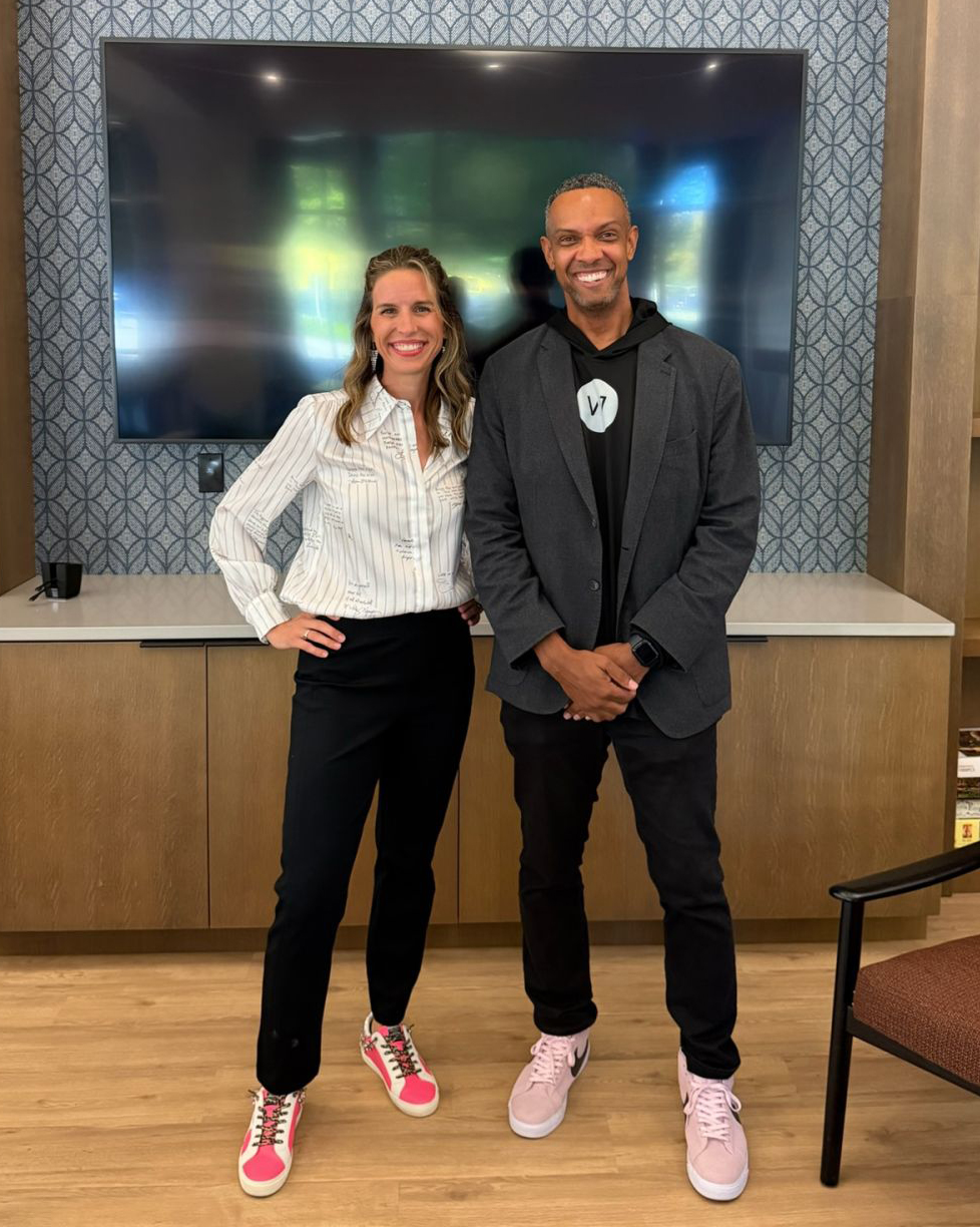 Two colleagues, a woman and a man, smiling and standing together in front of a wall-mounted TV screen at an office or event space. Two colleagues, a woman and a man, smiling and standing together in front of a wall-mounted TV screen at an office or event space.