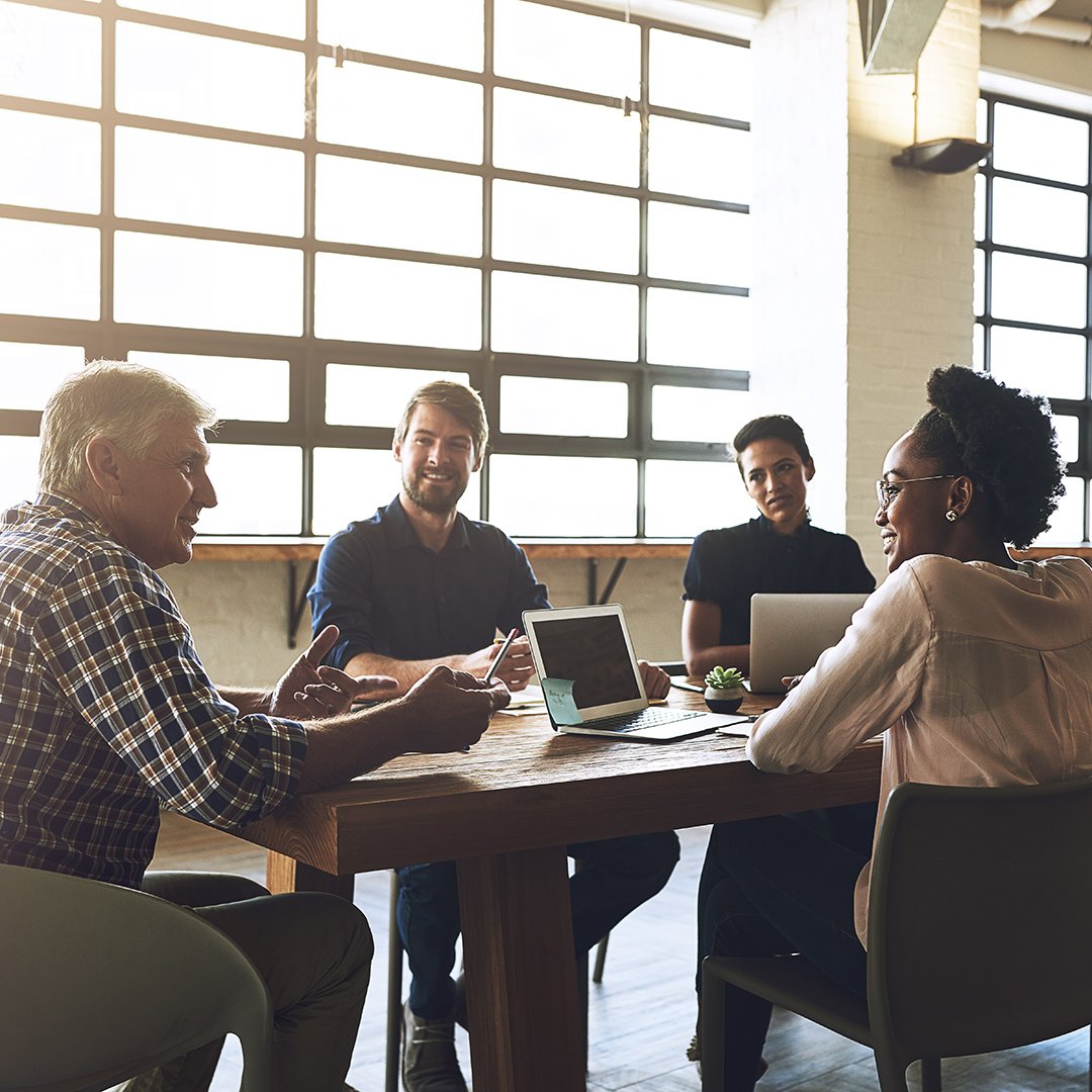 Four colleagues sit around a wooden table with laptops, having a discussion in a sunlit office with large industrial windows. Four colleagues sit around a wooden table with laptops, having a discussion in a sunlit office with large industrial windows.