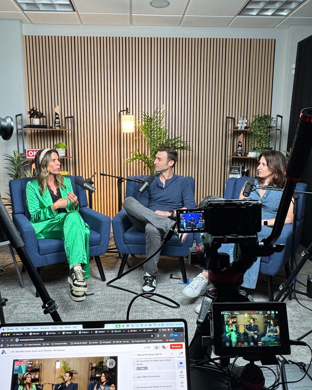Three people seated in blue armchairs recording a podcast in a studio, with cameras, microphones, and a laptop showing the livestream in the foreground.