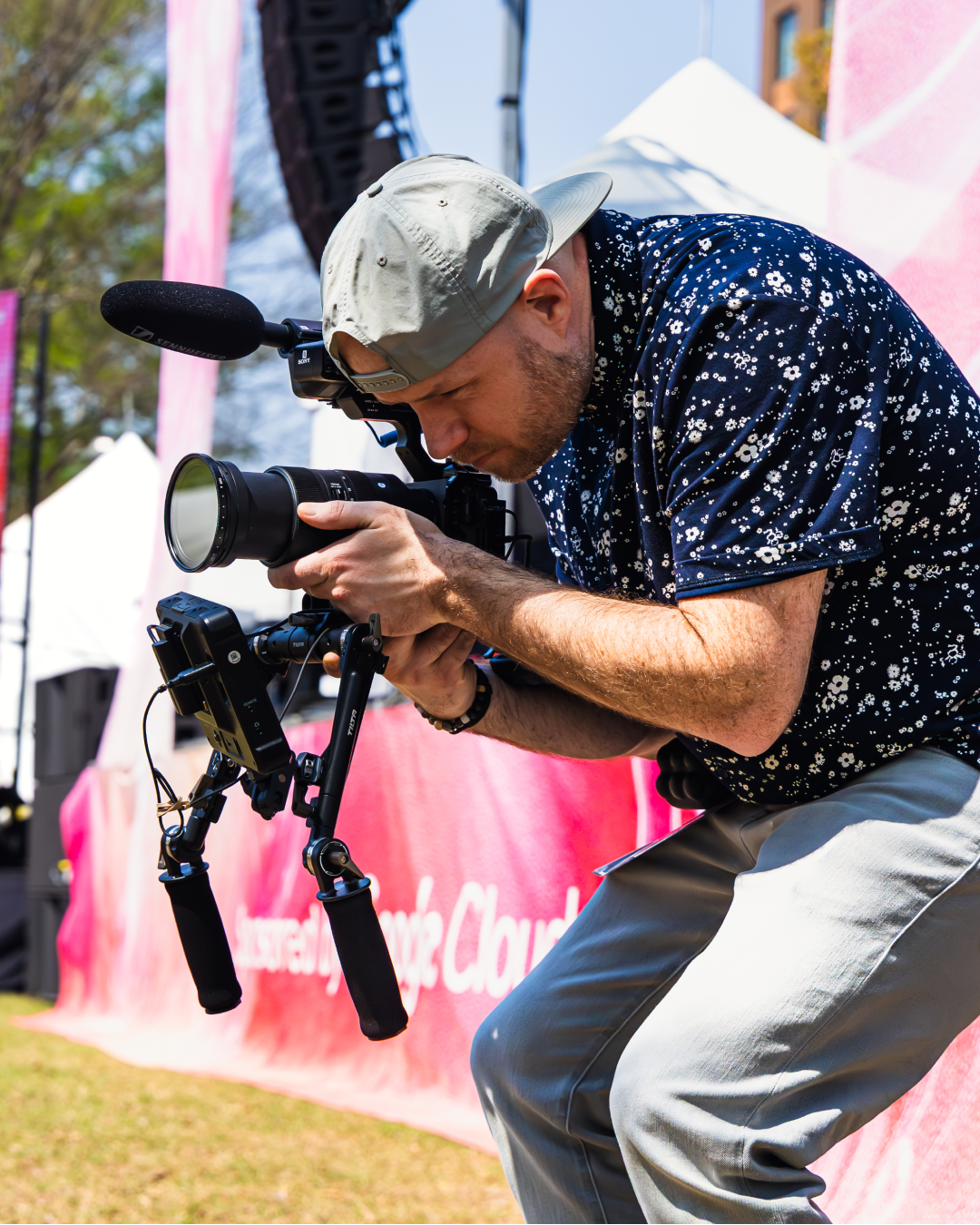 A videographer in a floral shirt and cap crouches while filming with a professional camera rig at an outdoor event.