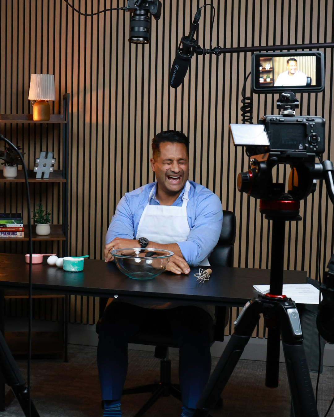 A man in a blue shirt and white apron laughs while sitting at a desk with baking ingredients, being filmed by multiple cameras.
